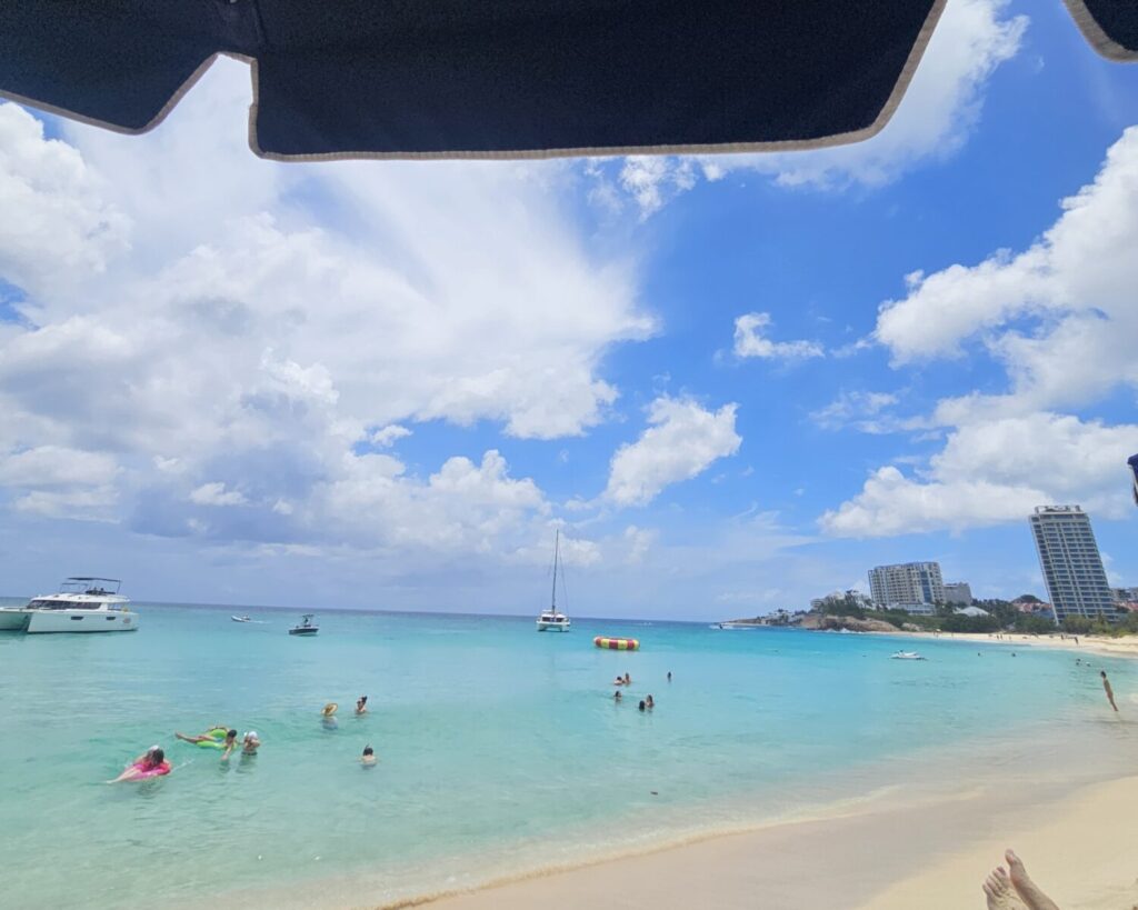 People swim and relax in clear blue ocean water near boats and a sandy beach, with buildings visible in the background.