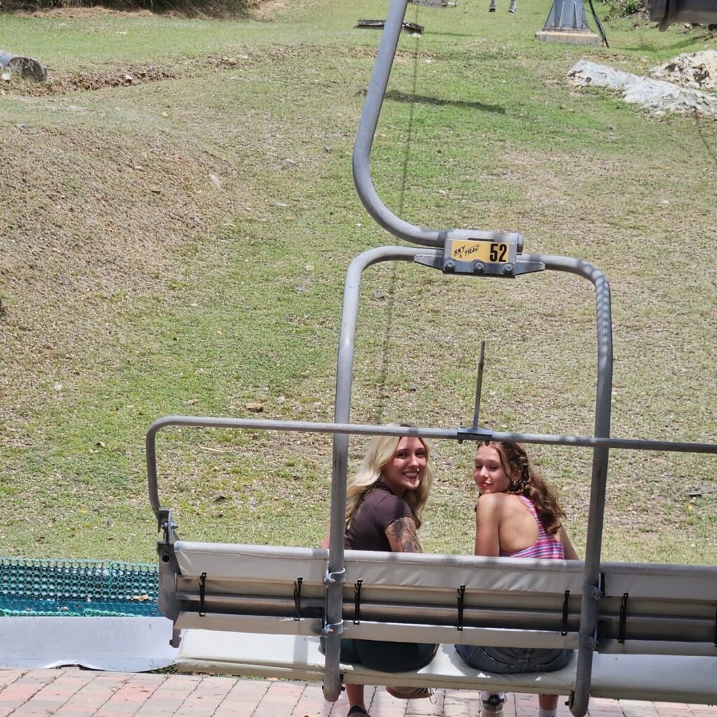 Two women are seated on a chairlift, facing backward, with grassy terrain visible in the background.