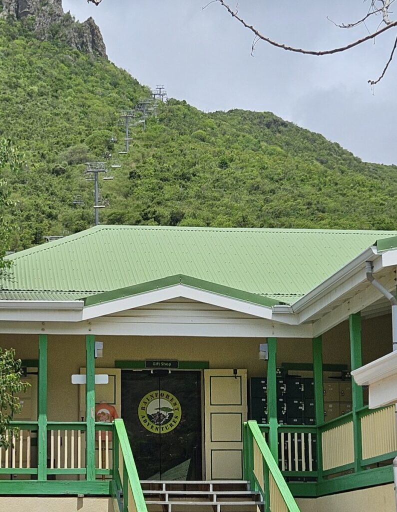 Green-roofed building with a gift shop sign in front of a steep, green hillside with a cable car line.