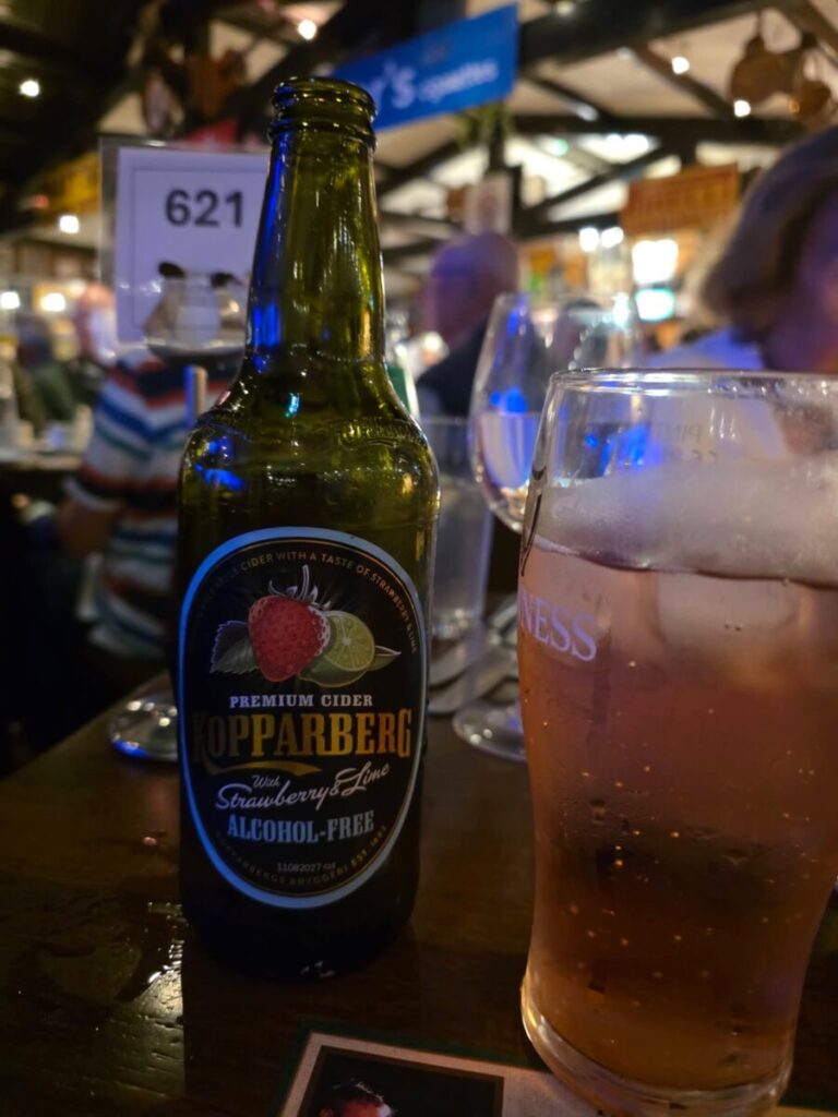A bottle of Kopparberg strawberry and lime alcohol-free cider next to a pint glass on a pub table.