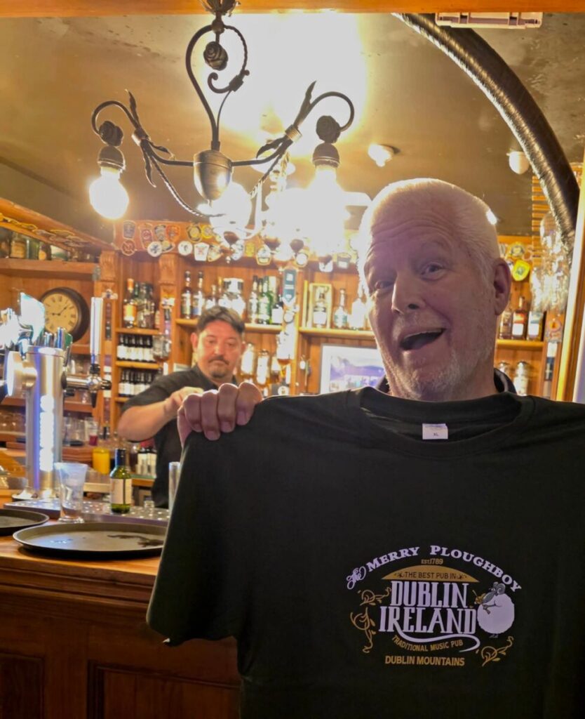 A man holds up a "Merry Ploughboy Dublin Ireland" T-shirt in a pub, with a bartender working behind the bar.