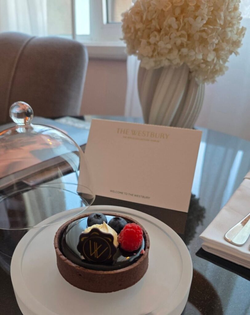 A chocolate tart with berries on a table beside a card reading “The Westbury” and a vase of white flowers.