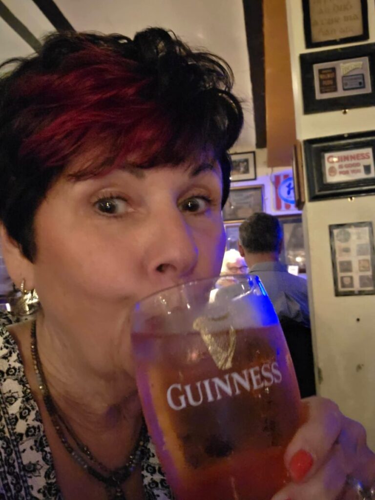 A person with short, dark hair drinks from a Guinness glass in a dimly lit bar decorated with framed pictures.